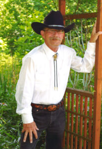 A man in a cowboy hat and white shirt stands by a wooden trellis, surrounded by greenery. He has a mustache and glasses.